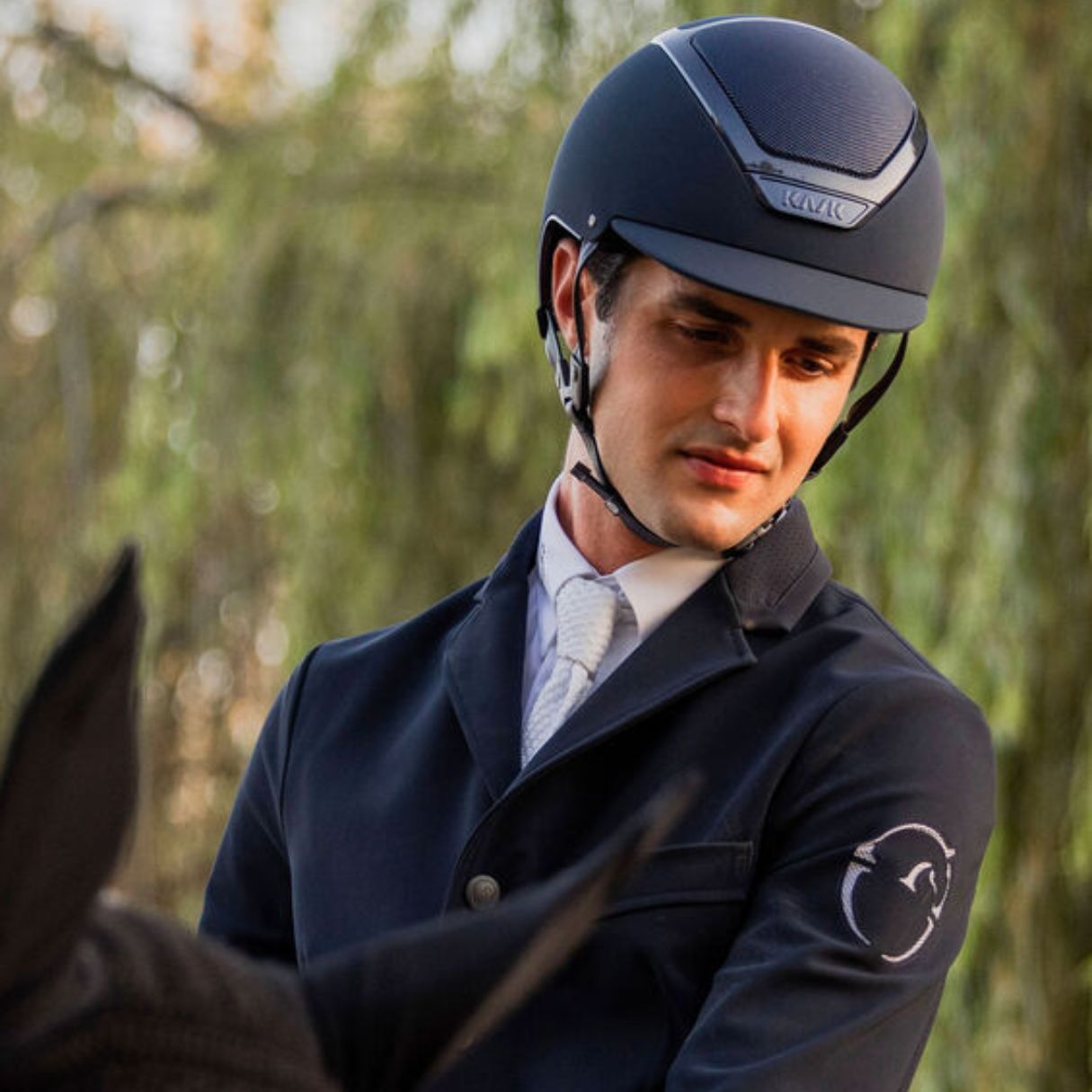 Kask Dogma Chrome Equestrian Helmet - Navy; shown on rider wearing showcoat, blurred trees in background | Malvern Saddlery