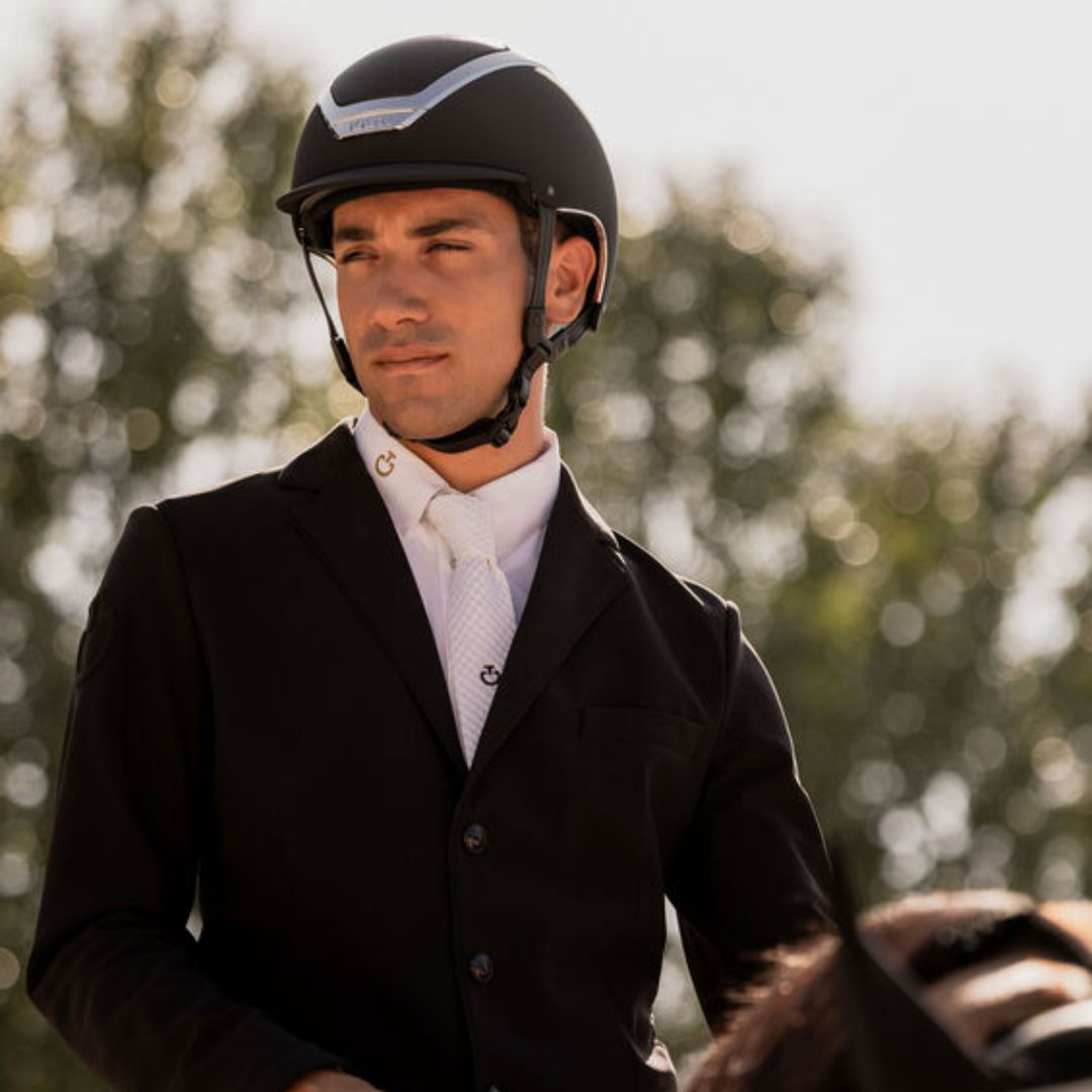 Man wearing a riding helmet and a black riding jacket, a white shirt, and a white tie, set against a blurred outdoor background of trees; Kask Dogma Chrome Equestrian Helmet - Black Matte with Silver Frame; lifestyle image | Standard Brim English Riding Helmets | Malvern Saddlery