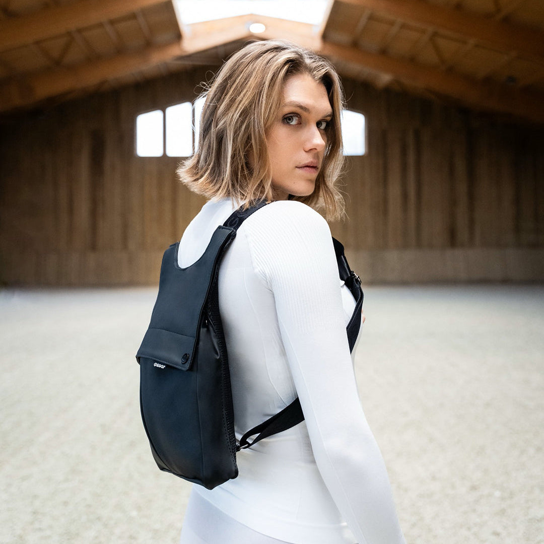 Equestrian rider wearing the black EGO7 Rosalie Backpack inside an indoor arena, featuring adjustable chest and shoulder straps over a white top