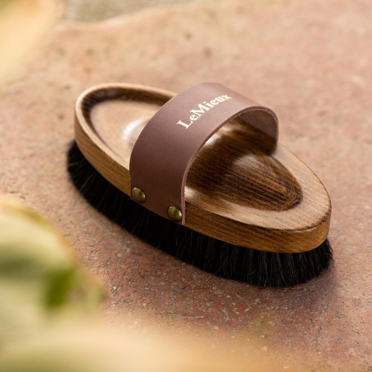 LeMieux Artisan Lambswool Body Brush - Brown with black bristles; shown on brick surface with blurred greenery in foreground | Horse Grooming | Malvern Saddlery
