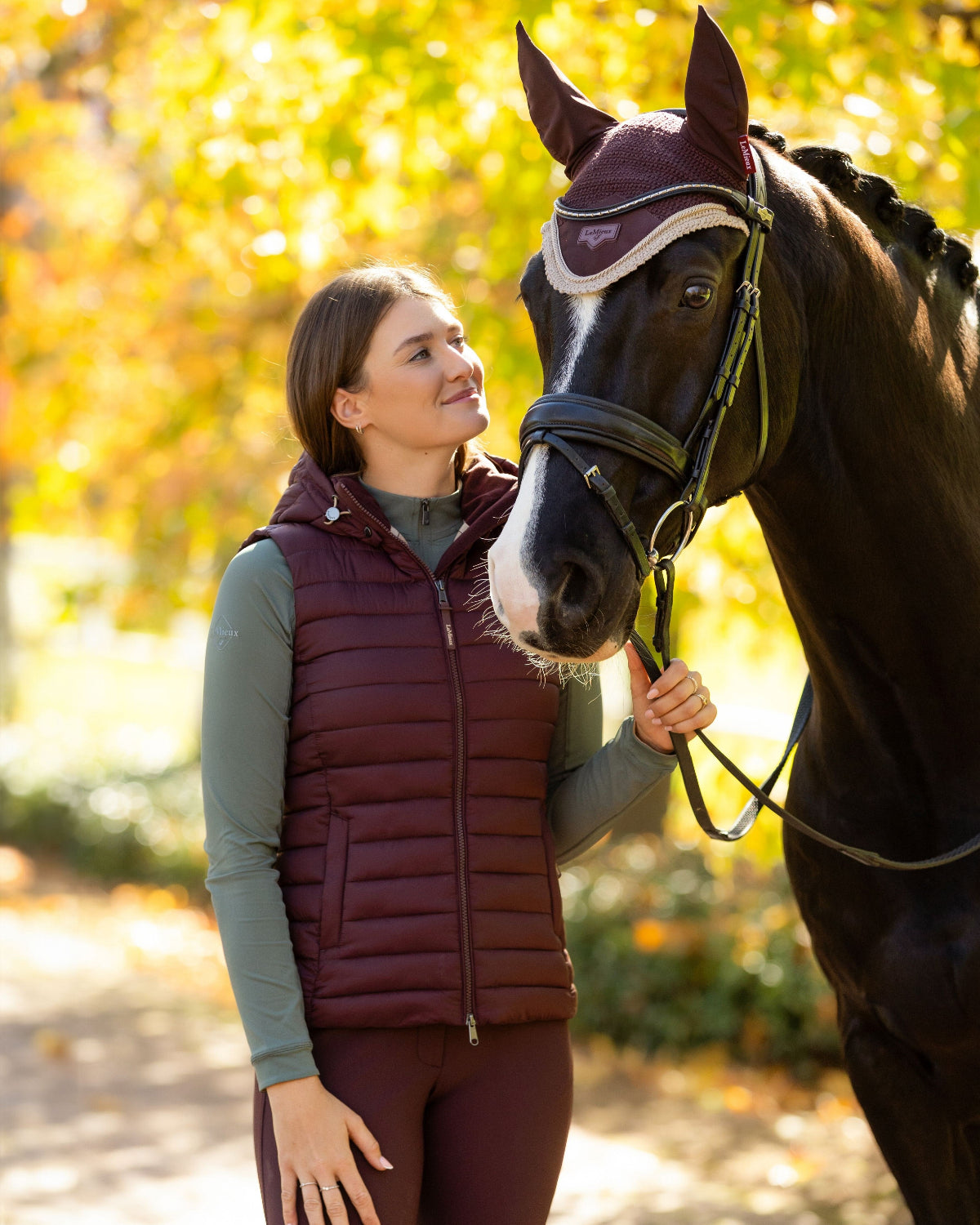 LeMieux Rose Hooded Puffer Vest - Damson Maroon, shown on woman standing next to horse with trees in background | Malvern Saddlery