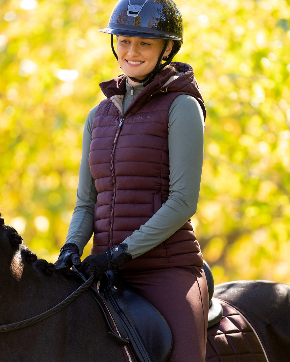 LeMieux Rose Hooded Puffer Vest - Damson Maroon, shown on woman riding horse with trees in background | Malvern Saddlery