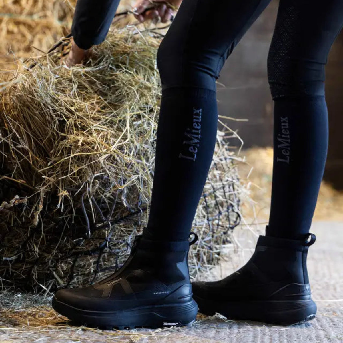 Equestrian wearing LeMieux Cavara Performance High Top Trainers in black while working near hay bales at the stable.