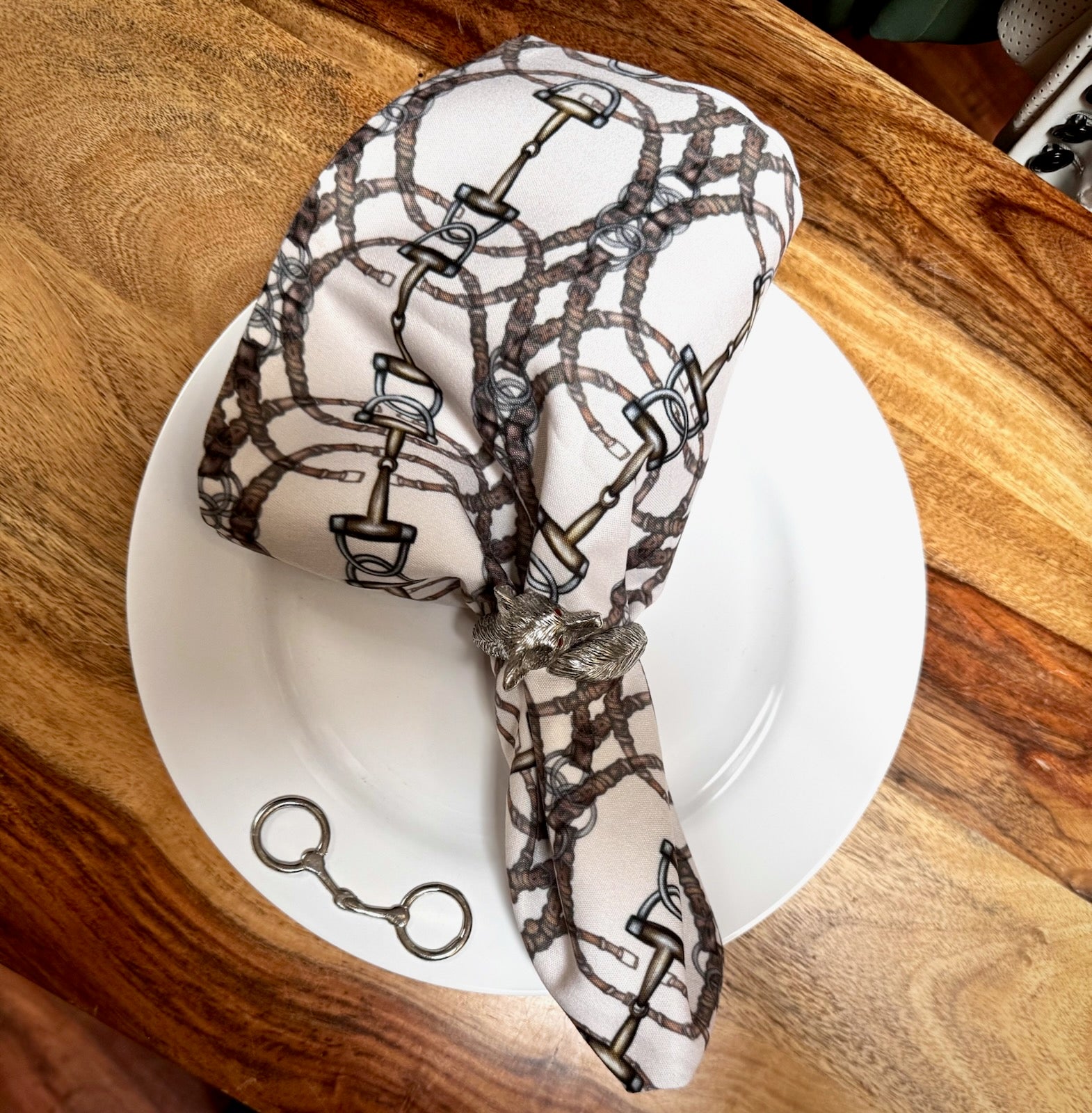 Equestrian Snaffle Bit Status Print Cloth Napkin, shown on white plate with bit accent & pewter fox napkin ring | Malvern Saddlery