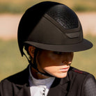 A close-up of a person wearing a black KASK equestrian Black Star Lady riding helmet with a mesh ventilation panel and a visor, set against a blurred outdoor background | Malvern Saddlery