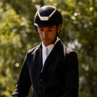 Kask Dogma Chrome Equestrian Helmet - Black Matte with Gold Frame; shown on man wearing equestrian show attire against blurred tree background | Standard Brim English Riding Helmets | Malvern Saddlery