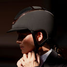 A close-up side profile of a person wearing a matte black equestrian riding helmet with a visor and black chin strap, set against a dark, shadowed background. Kask Dogma Chrome Equestrian Helmet - Black Matte with Rose Gold Frame | Standard Brim English Riding Helmets | Malvern Saddlery