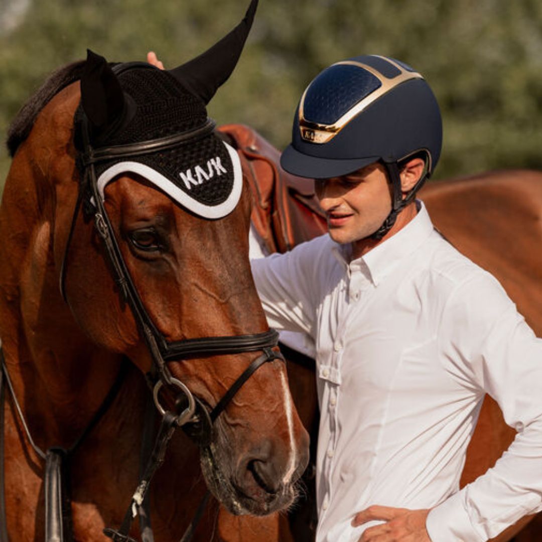 A person in a white shirt and Kask Dogma Chrome Equestrian Helmet - Navy Matte with Gold Frame;   stands beside a brown horse ; Standard Brim English Riding Helmets | Malvern Saddlery
