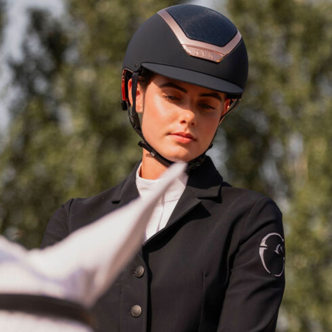 A woman in equestrian attire wearing a Kask Dogma Chrome Equestrian Helmet - Navy Matte with Rose Gold Frame;   and a black riding jacket with a white shirt underneath, set against a blurred outdoor background of trees | Standard Brim English Riding Helmets | Malvern Saddlery