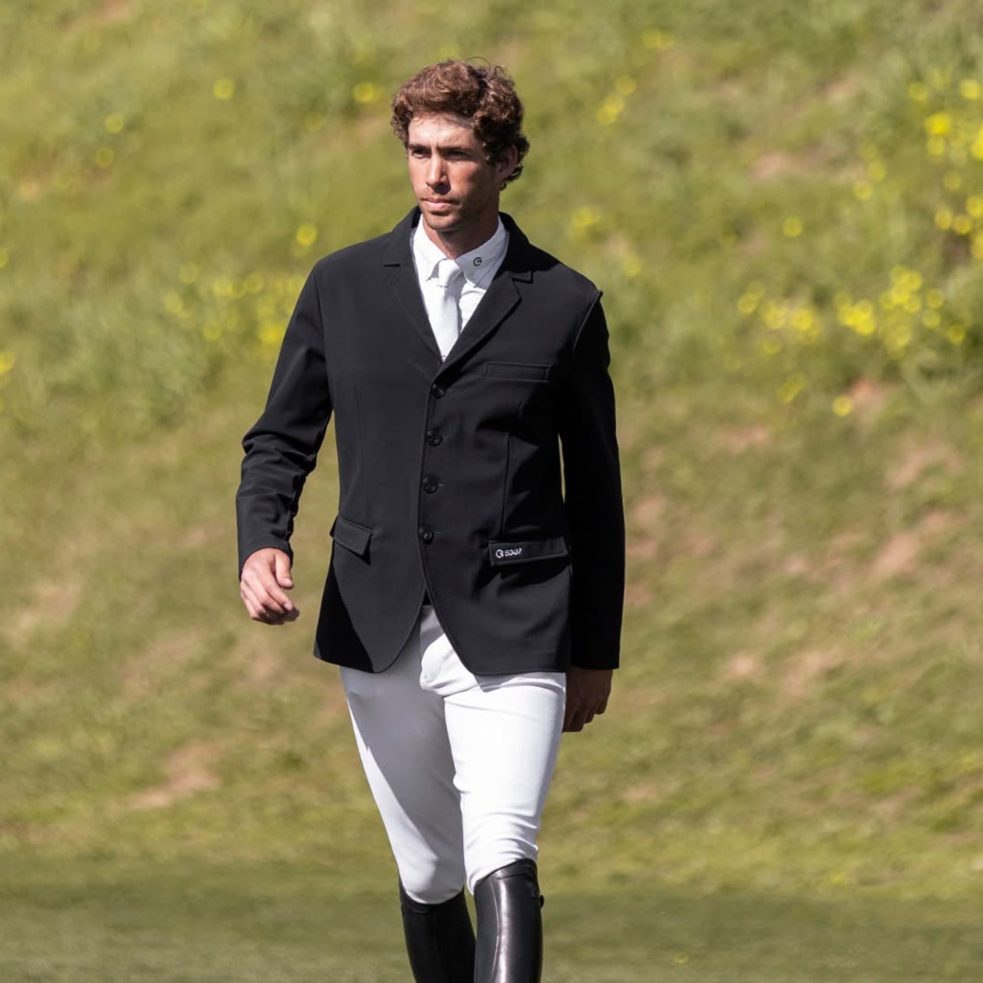 Male rider wearing the black EGO7 Elegance Men's Show Jacket, white competition breeches, and black tall boots, walking outdoors across a grassy field.