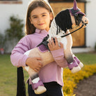 Young girl holding a LeMieux Toy Pony Toby [with tack & accessories] outdoors with a building and plants in the background | Malvern Saddlery