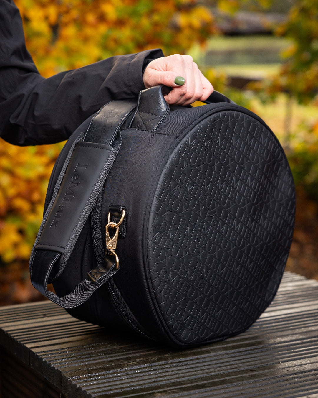 LeMieux Luxe Hat/Helmet Box - Black , shown outdoors on table with trees in background woman's hand on handle | Rider Travel Accessories | Malvern Saddlery
