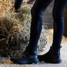 Equestrian wearing LeMieux Cavara Performance High Top Trainers in black while working near hay bales at the stable.