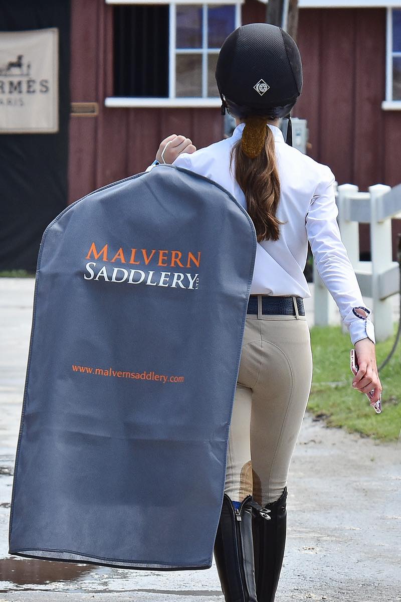 Equestrian rider with Malvern Saddlery garment bag walks toward barn